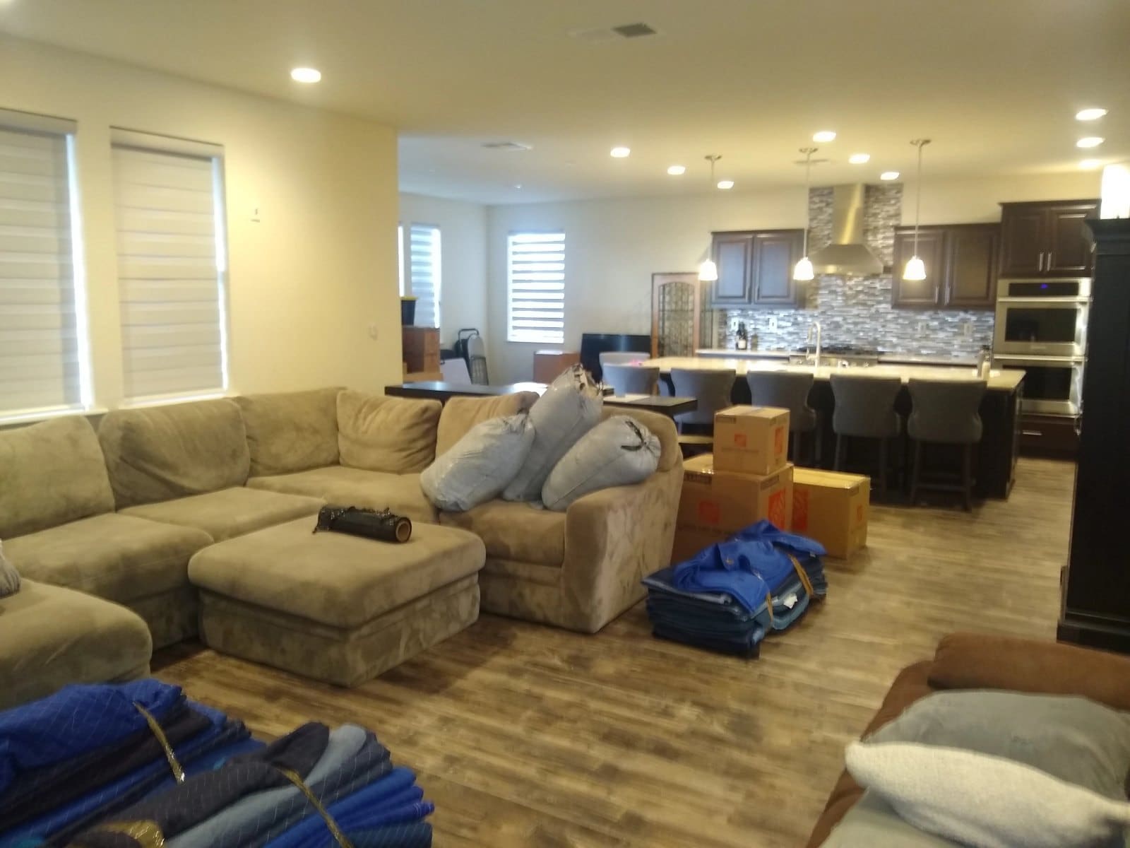 Open-concept living room with beige sectional sofa, cardboard boxes, and kitchen with dark cabinets visible in background