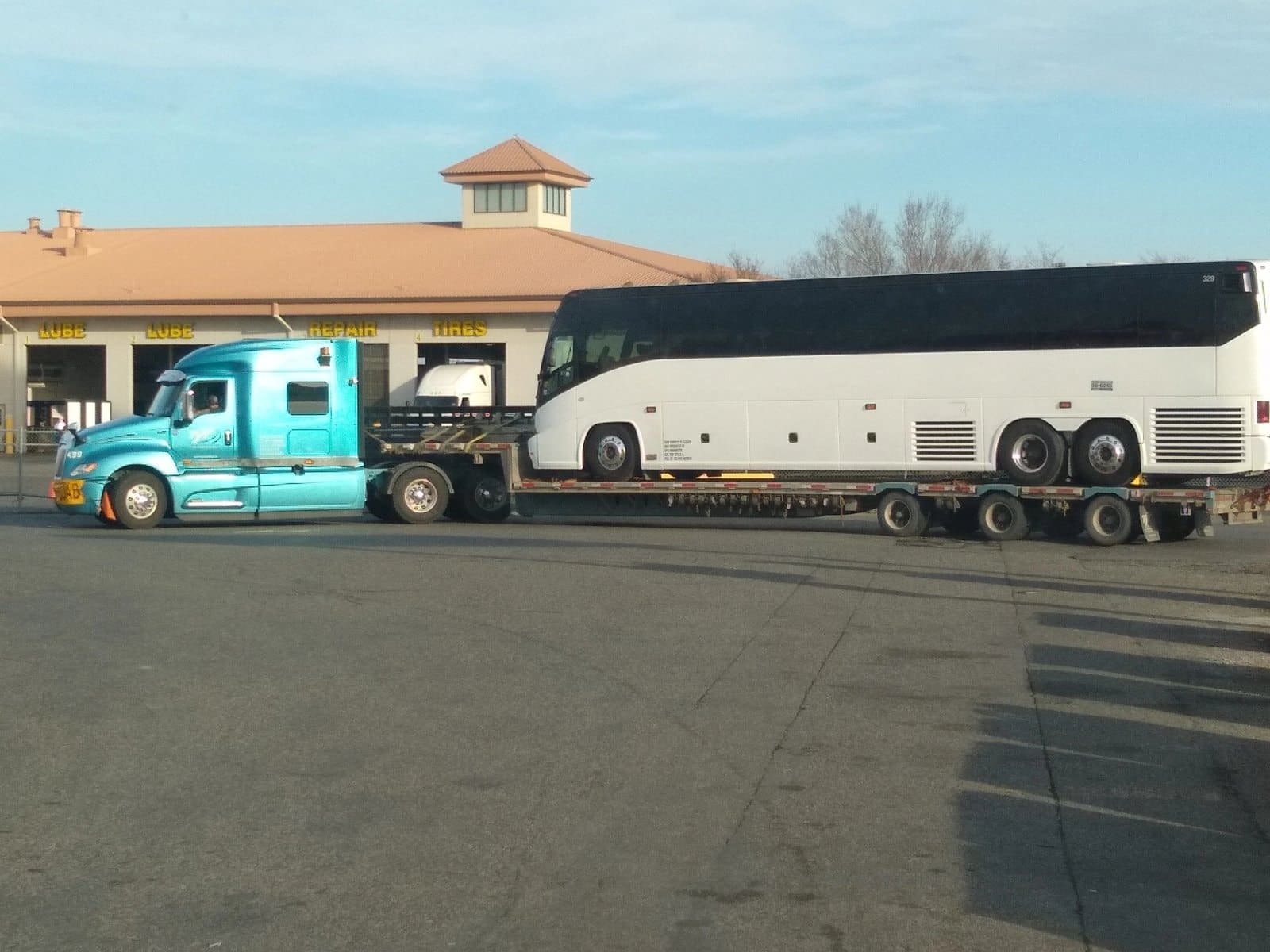 Turquoise semi truck hauling white tour bus on flatbed trailer at service station