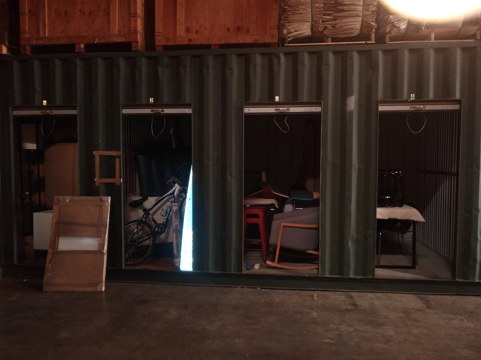 Four corrugated metal storage units with dark interiors containing furniture, a bicycle, and household items