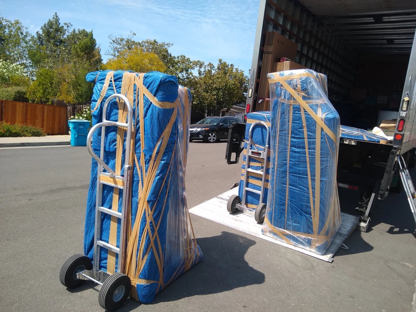 Two blue wrapped furniture items on hand trucks outside a moving truck