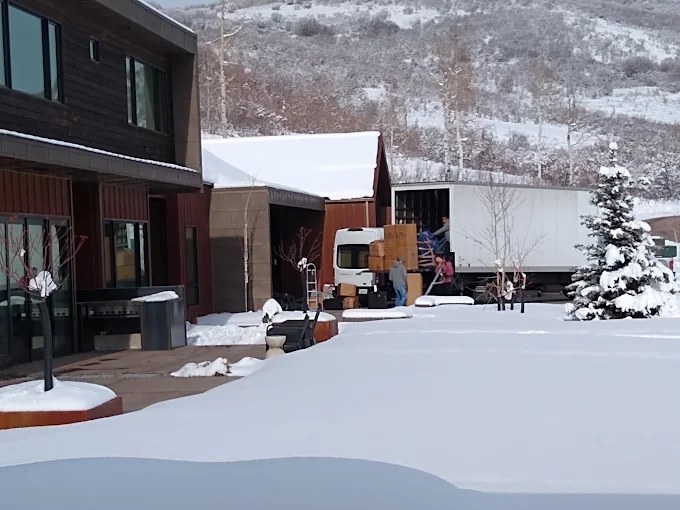 Moving trucks and workers unloading boxes at snowy commercial building with mountains in background