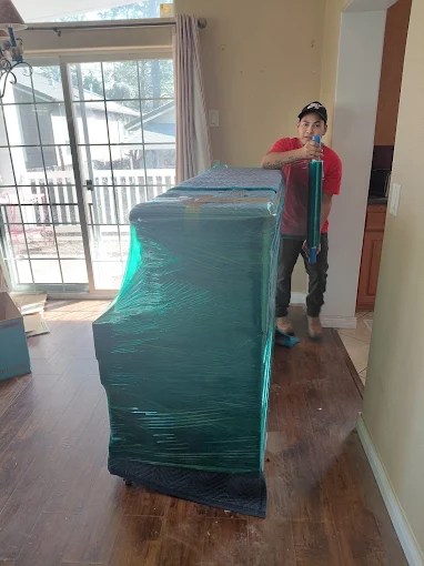 Young person wrapping large appliance in green plastic wrap in apartment hallway
