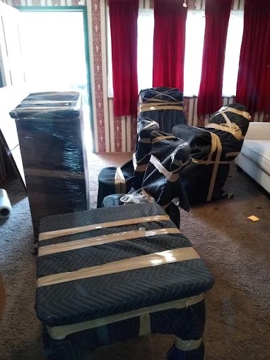 Furniture wrapped in plastic and strapped with moving bands stacked in an empty room with burgundy curtains
