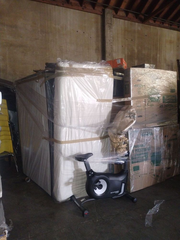 Stationary exercise bike wrapped in plastic in warehouse storage area surrounded by boxed items